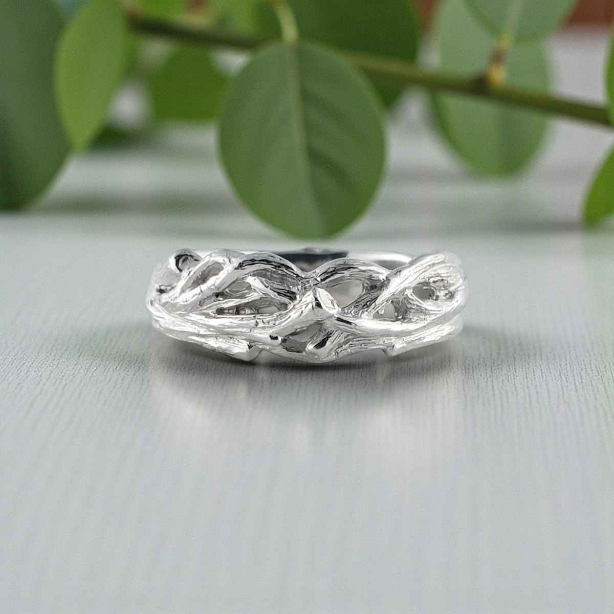 white gold ring with leaf design on a light gray surface with green leaves in the background