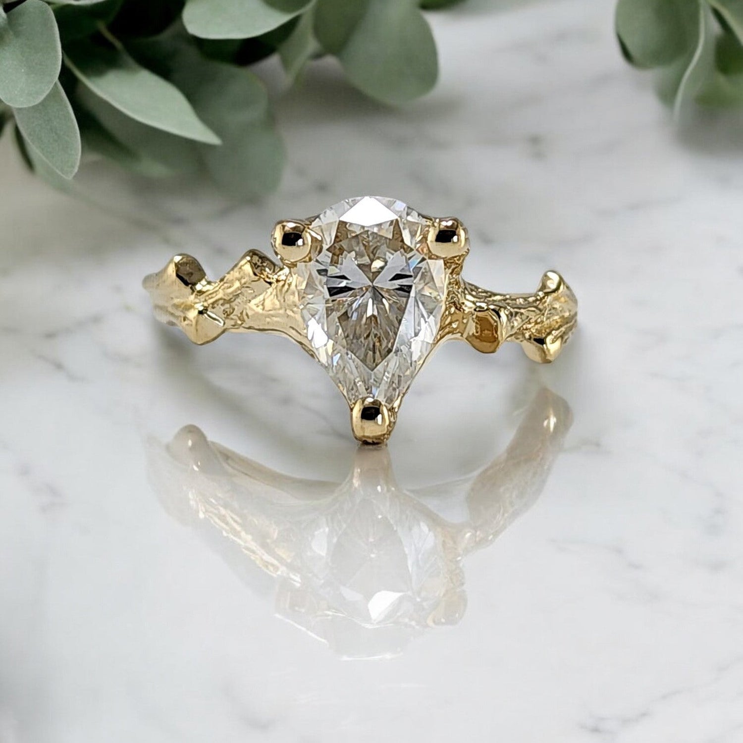 pear shaped moissanite in a yellow gold twig style ring sitting on a marble table